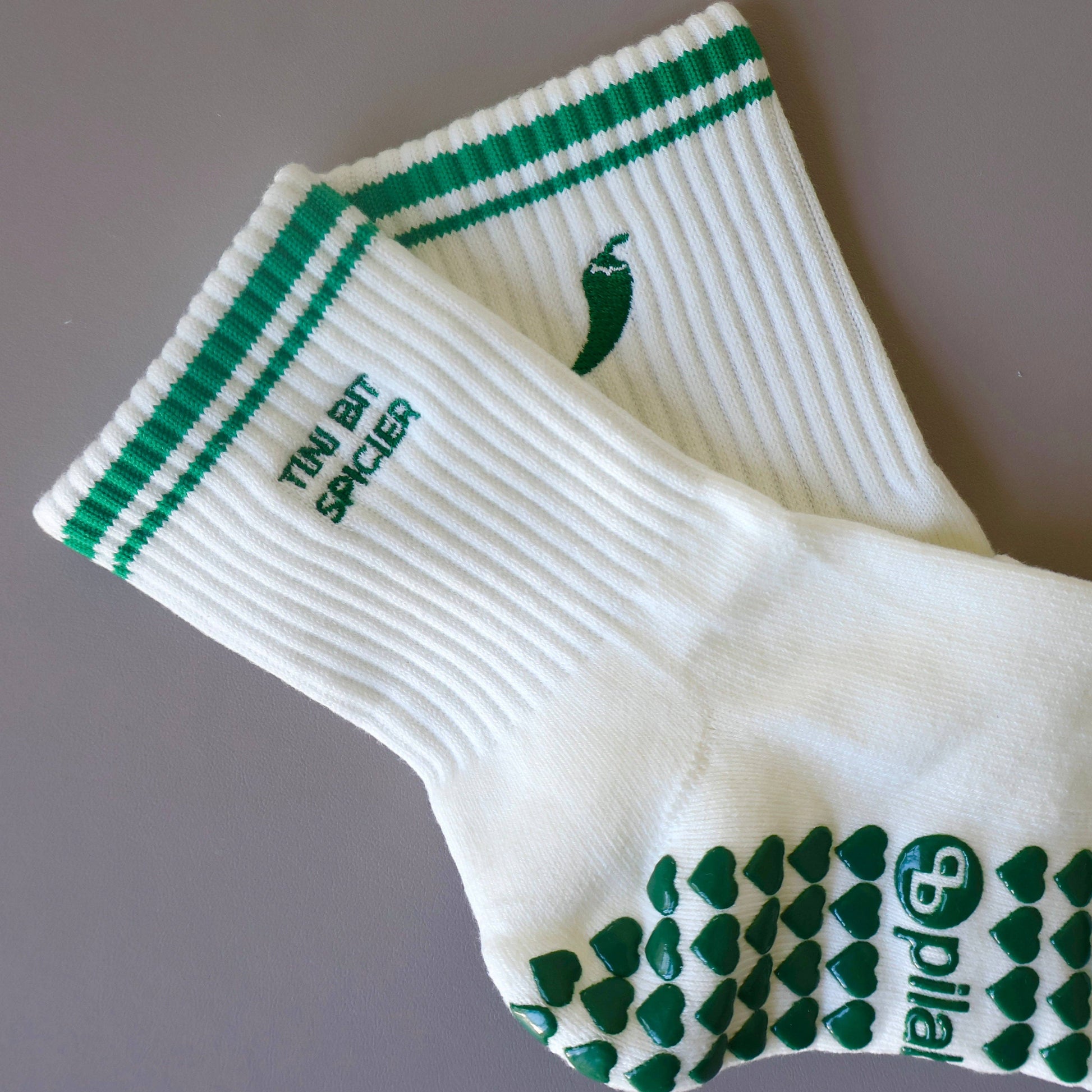 White and green Pilates grip socks with striped ribbed cuffs, embroidered chili pepper detail and green non-slip grips on the soles.