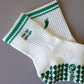 White and green Pilates grip socks with striped ribbed cuffs, embroidered chili pepper detail and green non-slip grips on the soles.